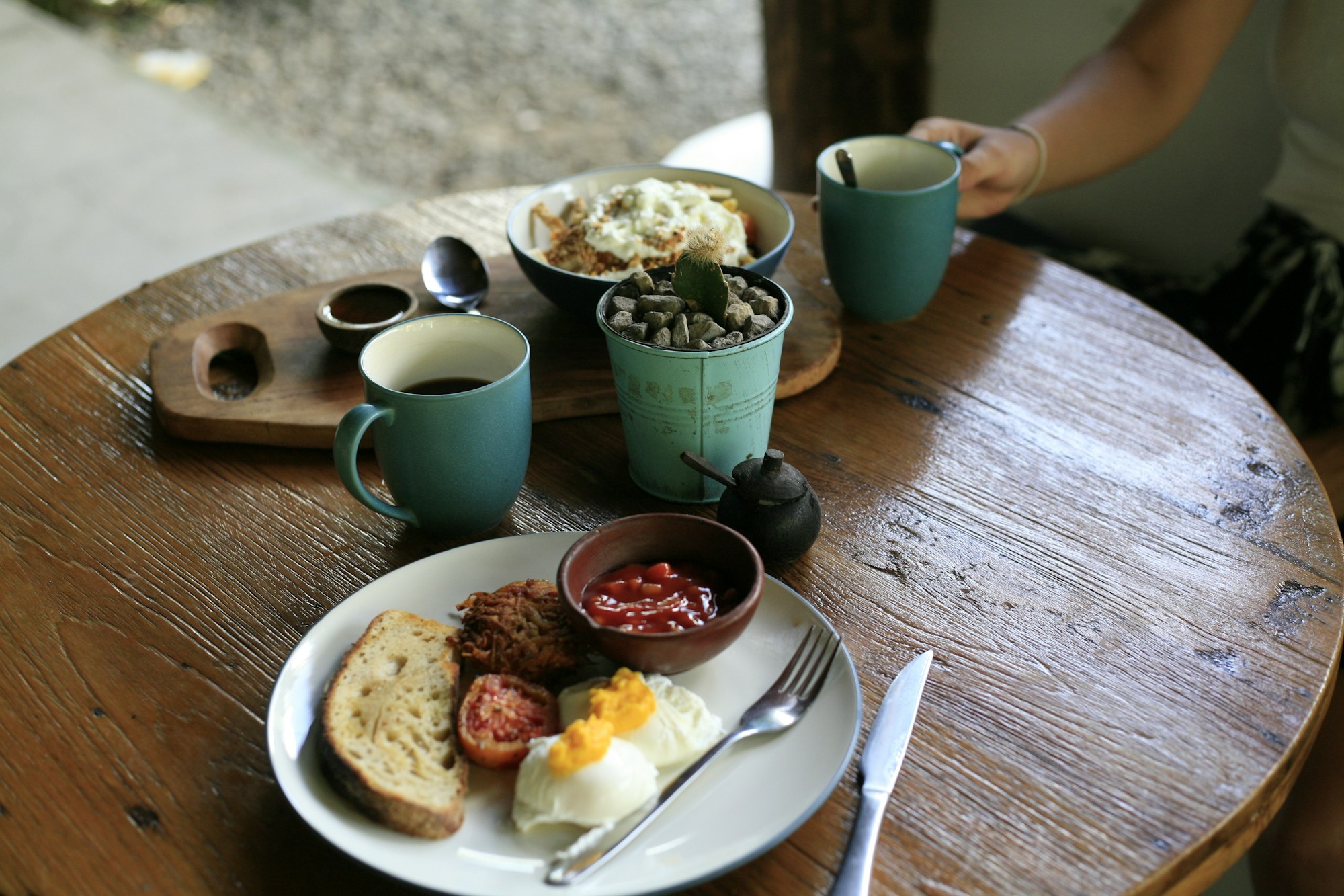 A cooked breakfast of poached eggs, sourdough toast and roast tomato on a round wooden table, with coffee in a green mug and a second plate being carried in.