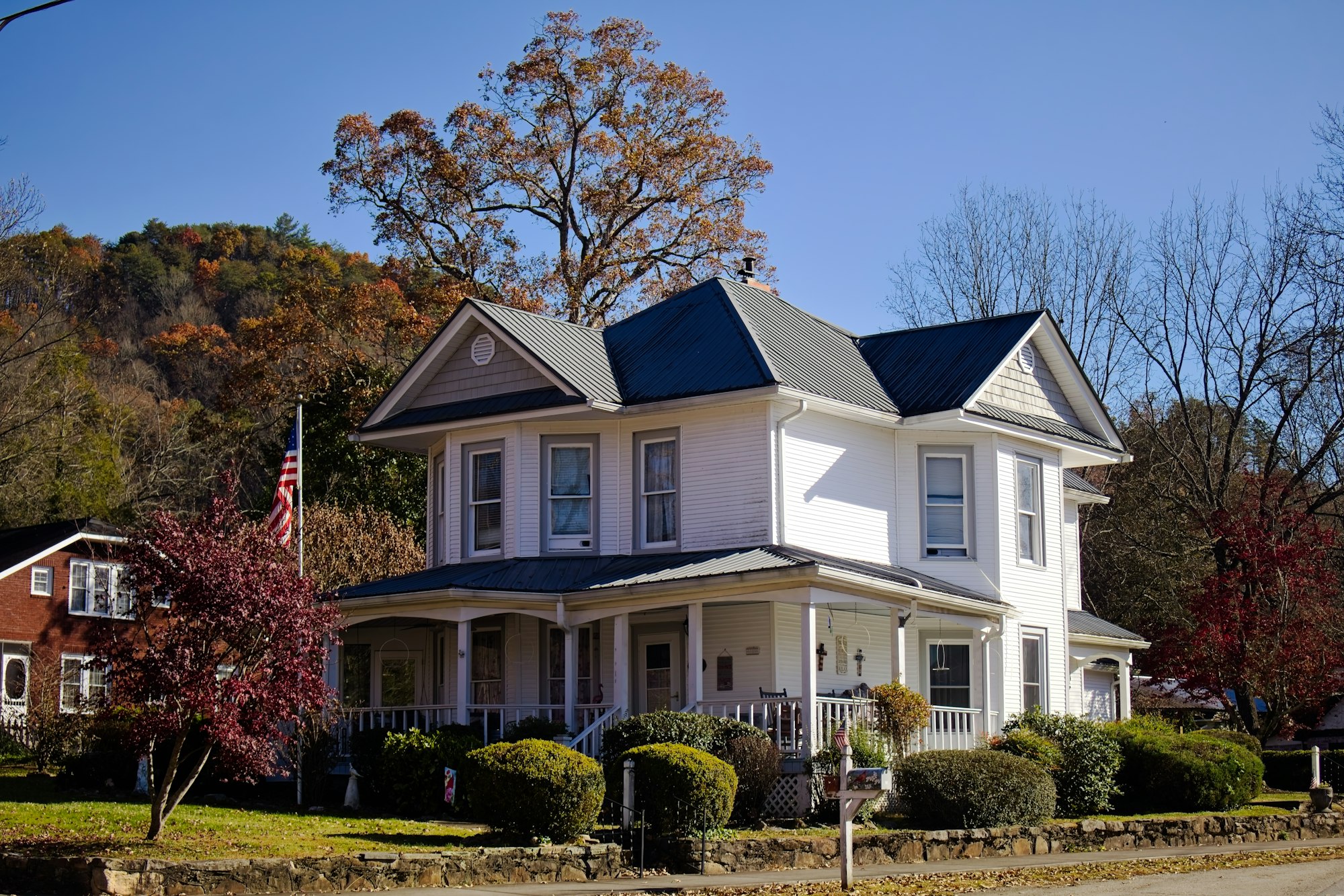 A white two storey clapboard house with a wraparound porch and a dark metal roof, photographed from the street in late autumn.