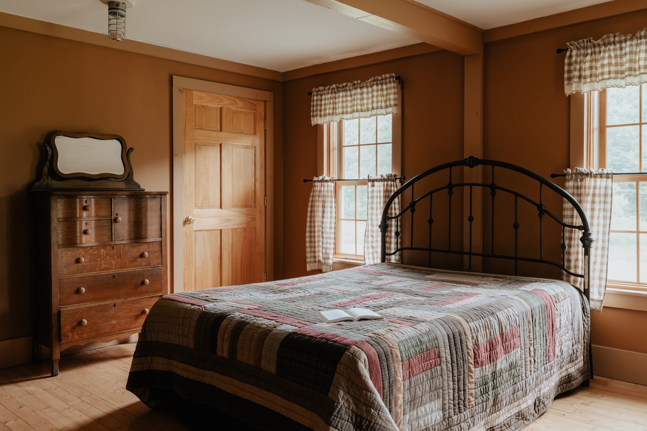 A bedroom in ochre paint with an iron bedstead under a patchwork quilt, an oak chest of drawers and two gingham curtained windows.