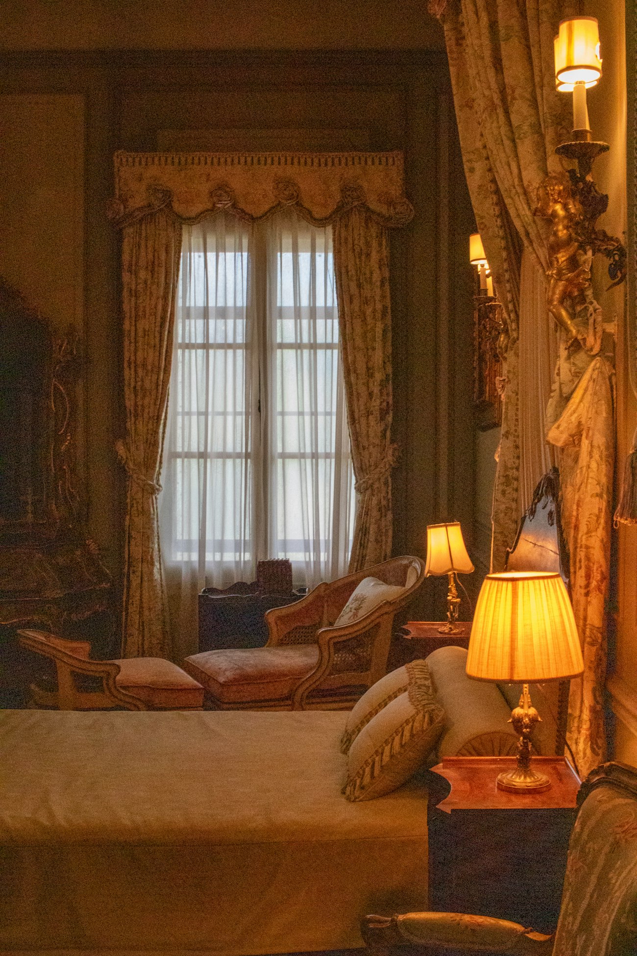A lamplit front bedroom with a tall curtained window, a velvet chaise under it and a brass lamp beside the bed.