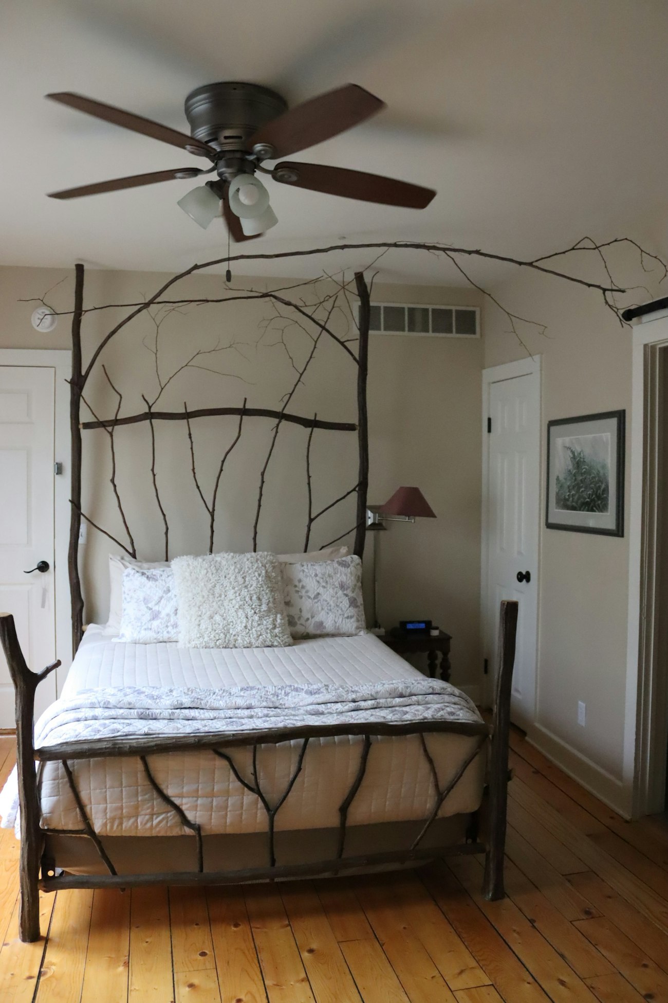 A plain ground floor bedroom with a bed framed in bare branches, white quilt, wide pine boards and a ceiling fan.