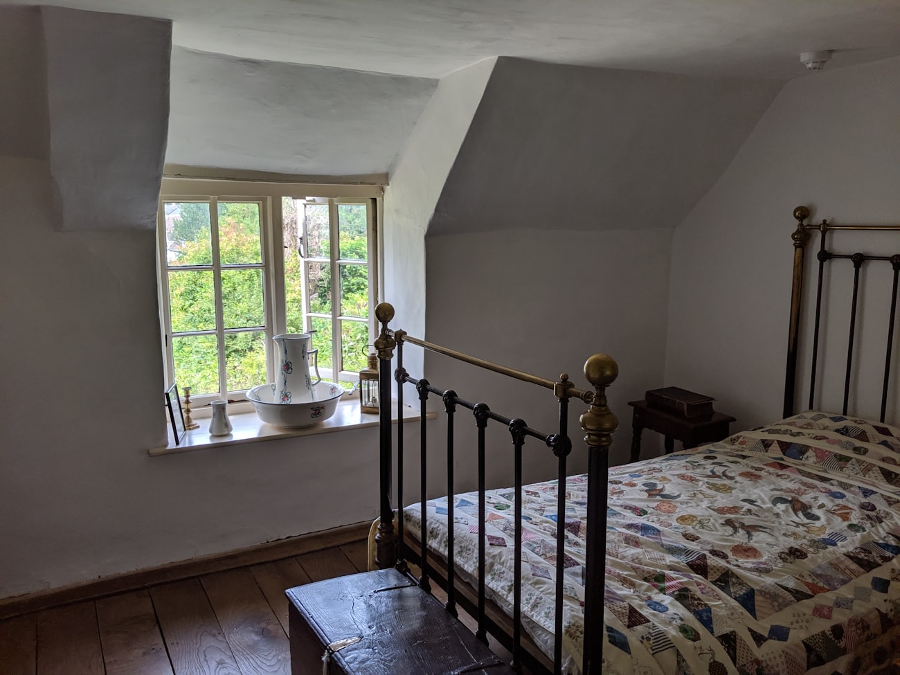 An attic bedroom under a sloping whitewashed ceiling, a brass and iron bed with a patchwork quilt, and a small window with a jug and basin on the sill.