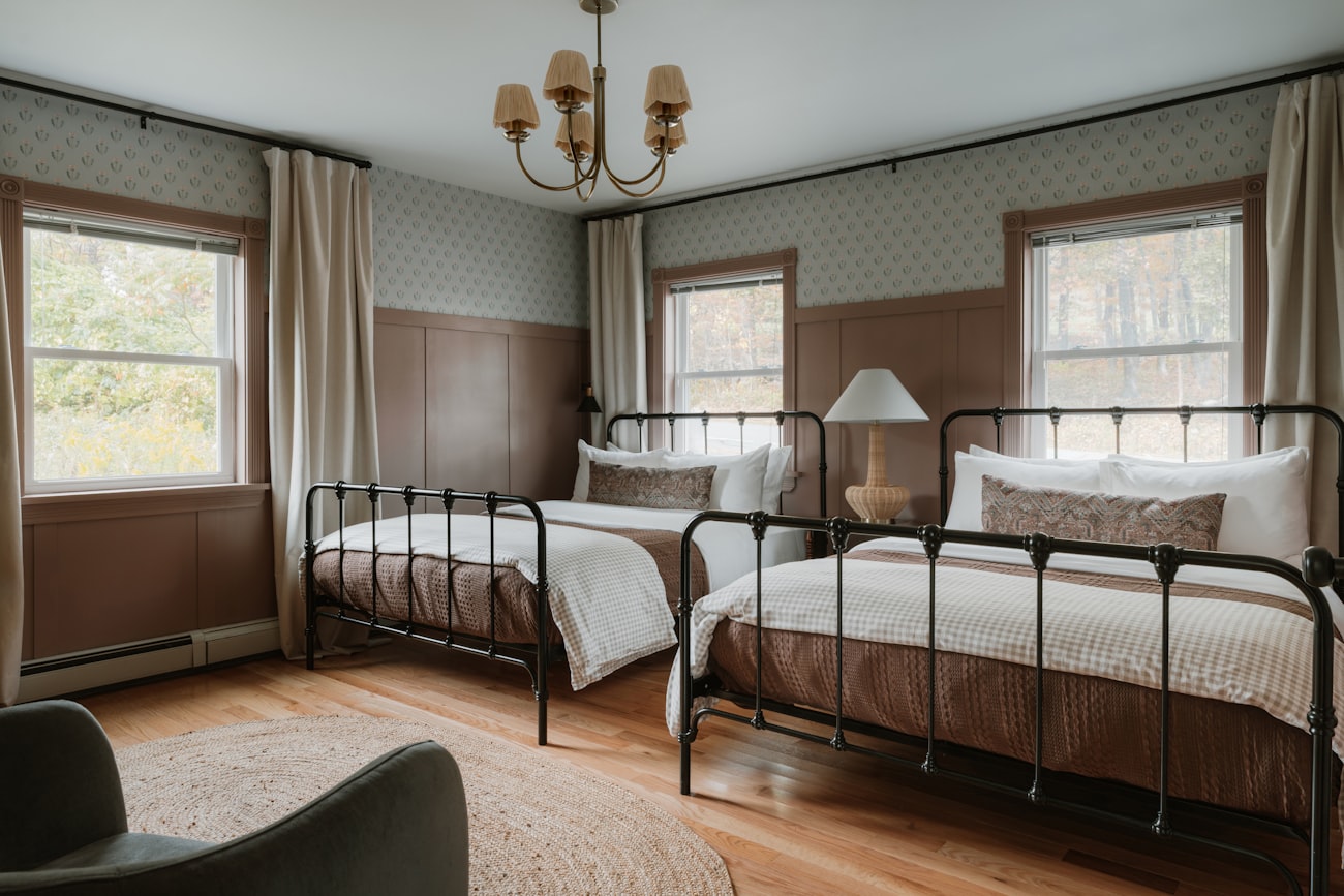A back bedroom with two black iron single beds side by side under patterned wallpaper, a rattan chandelier between them.